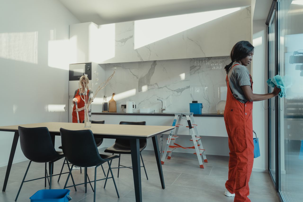services-01 Two women in work clothes cleaning a kitchen with glass doors.